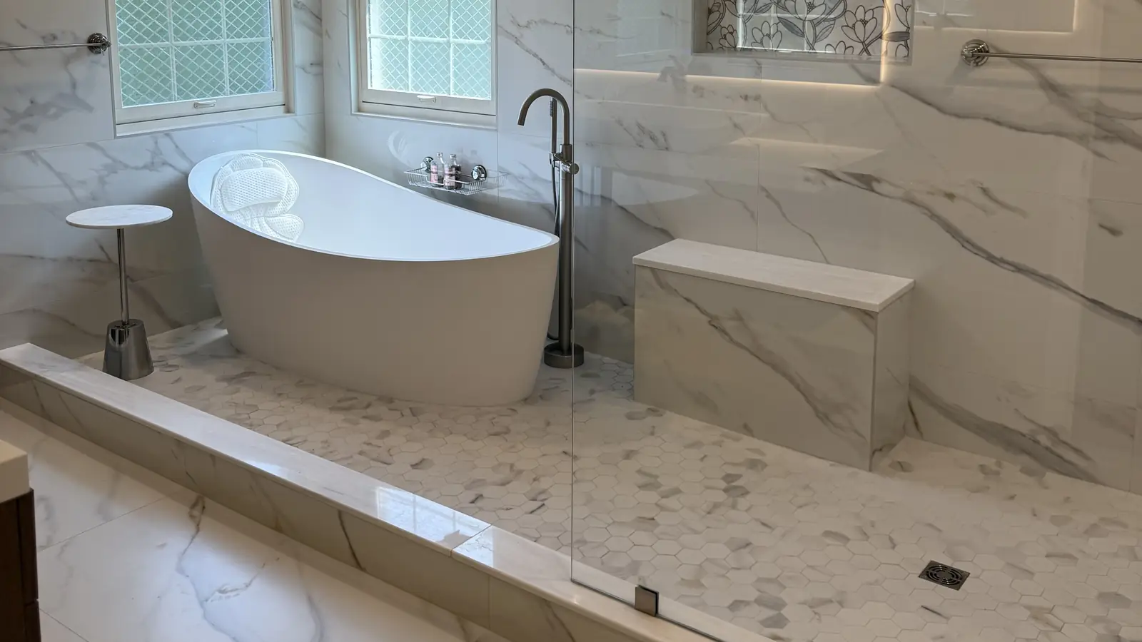 Primary bathroom in Burlington County, NJ: freestanding tub under two windows, marble-look porcelain walls and a walk-in shower with a bench